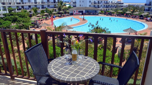 A poolside view with a round table and chairs, a pool, and a building with balconies in the background.