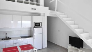 Een moderne keuken met witte kasten, een magnetron en een koelkast. Er is een eethoek met een rode tafel en stoelen. Een trap leidt naar een verdieping.
