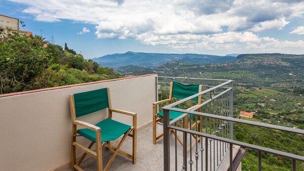 Een balkon met twee houten stoelen en een metalen hekwerk met uitzicht op een mooi landschap met heuvels en een verre berg.