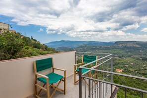 Un balcone con due sedie di legno e un parapetto metallico affacciato su un paesaggio pittoresco con colline e una montagna in lontananza.