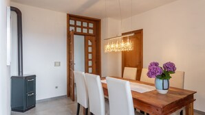 Een eetruimte met een houten tafel, witte stoelen, een kroonluchter en een vaas met paarse bloemen.