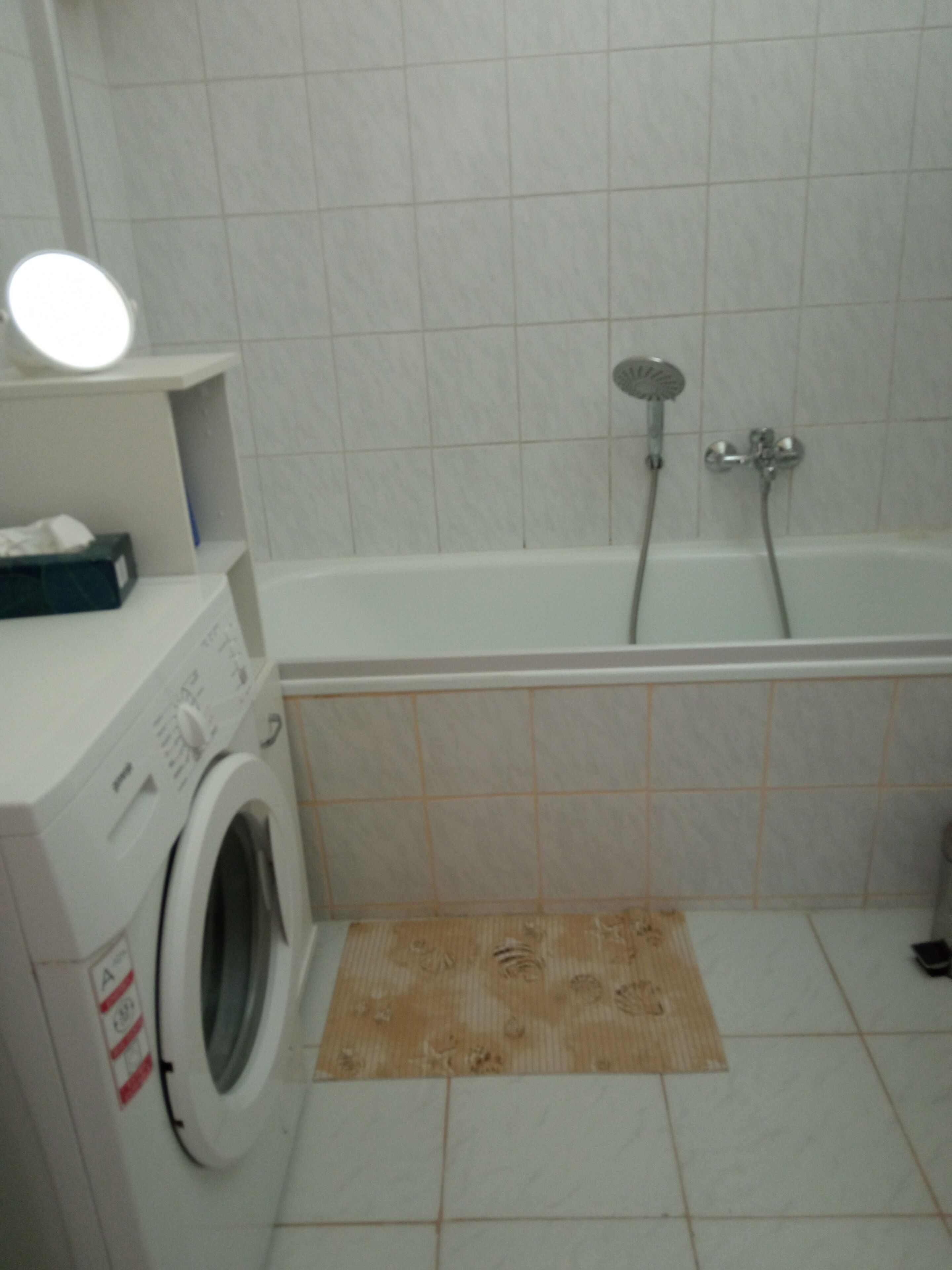 A bathroom with a washing machine, a bathtub with a showerhead, and a mirror on a shelf.