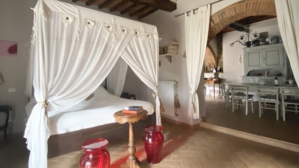 Une chambre avec un lit sous un dais, une petite table avec des livres et des vases rouges posés sur le sol.