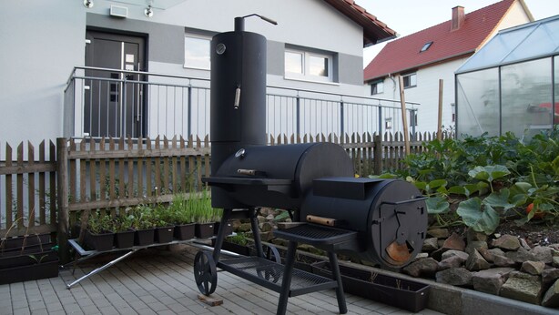 Ein schwarzer Grill mit Schornstein, ein Holzzaun und ein Garten mit großen, belaubten Pflanzen.
