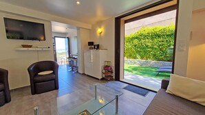 Een moderne woonkamer met een glazen salontafel, een bank, een aan de muur bevestigde televisie en uitzicht op een tuin door een schuifdeur.
