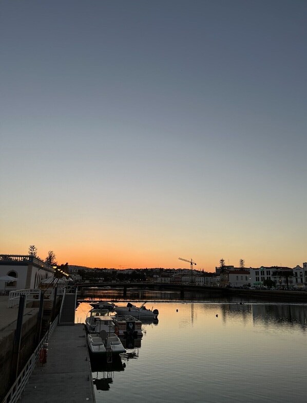 Un port de plaisance au bord de l’eau, au coucher du soleil, avec des bateaux amarrés, un pont en arrière-plan et des bâtiments le long de la rive.