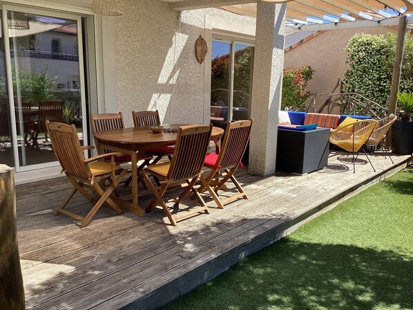 Une terrasse en bois avec une table ronde et des chaises, un espace salon couvert avec des coussins, et une vue sur le jardin environnant.