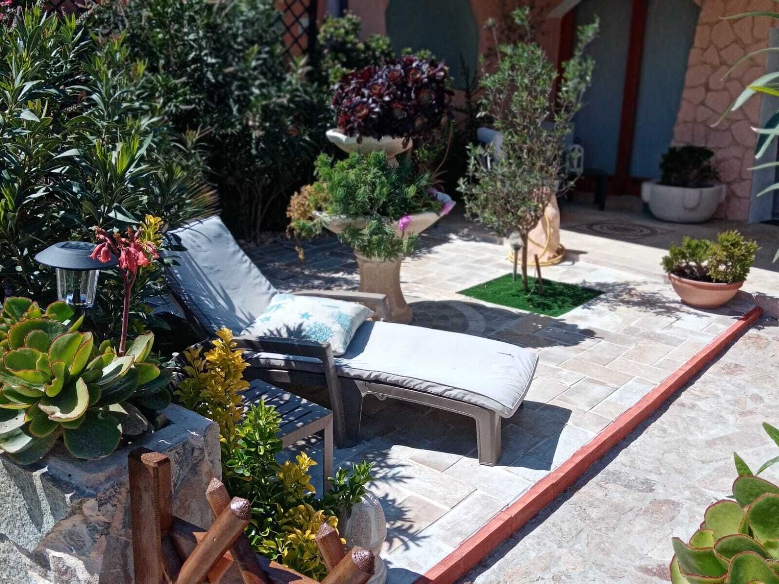 A sunlit patio with a lounge chair, various potted plants, and a stone wall.