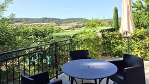 Eine Terrasse im Freien mit einem runden Tisch und Stühlen, einem beigen Sonnenschirm und Blick auf eine grüne Landschaft mit Bäumen und Hügeln.