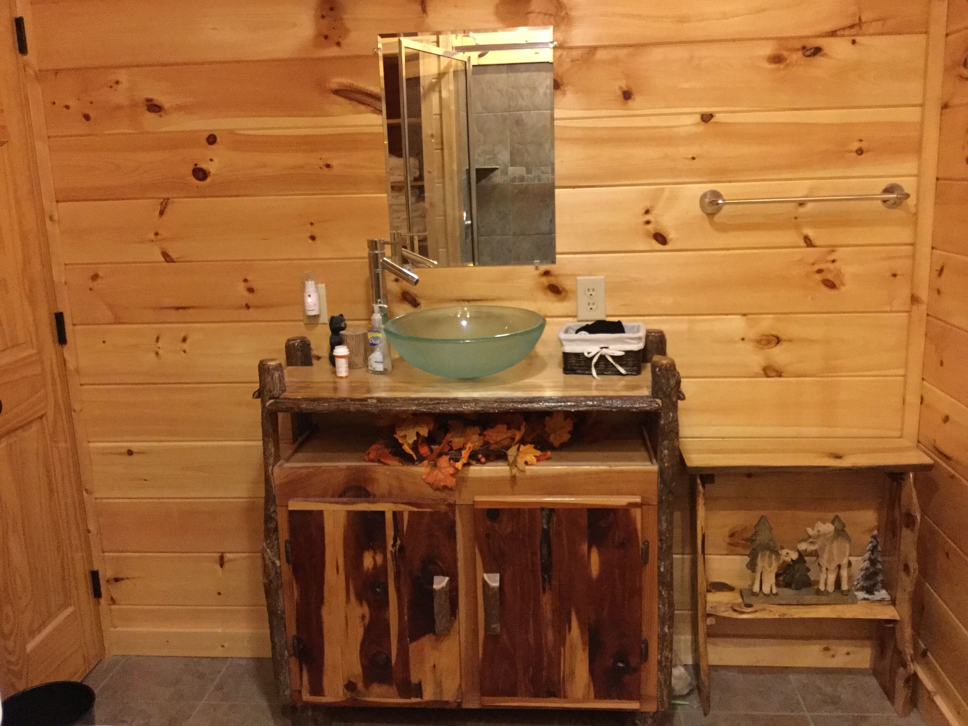 A wooden cabin bathroom with a glass bowl sink, a mirror, and a small shelf with toiletries.