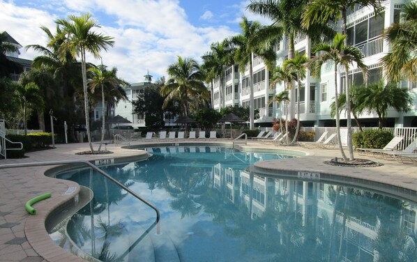 A swimming pool surrounded by palm trees and lounge chairs, with a multi-story building in the background.