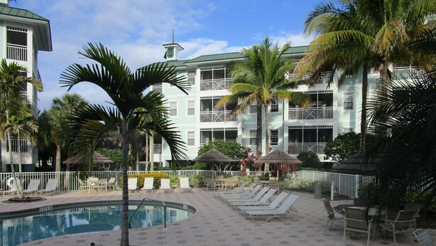 A pool area with lounge chairs, palm trees, and a building with balconies.