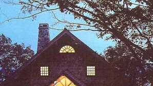 Una cabaña de troncos con techo inclinado, una chimenea central y ventanas dispuestas simétricamente.