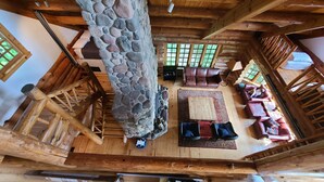 Interior rústico de una cabaña con chimenea de piedra, vigas de madera, una escalera de madera y una zona de estar acogedora con sofá y sillones.