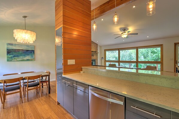 A modern kitchen with a large island, wooden paneling, stainless steel appliances, pendant lighting, and a view of greenery through large windows.