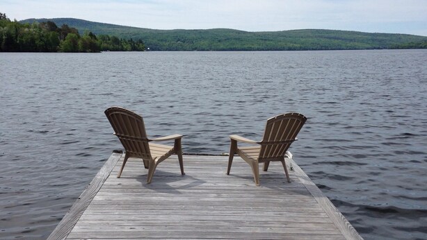Une jetée en bois avec deux fauteuils Adirondack donnant sur un lac paisible, le tout entouré de verdure et de collines.
