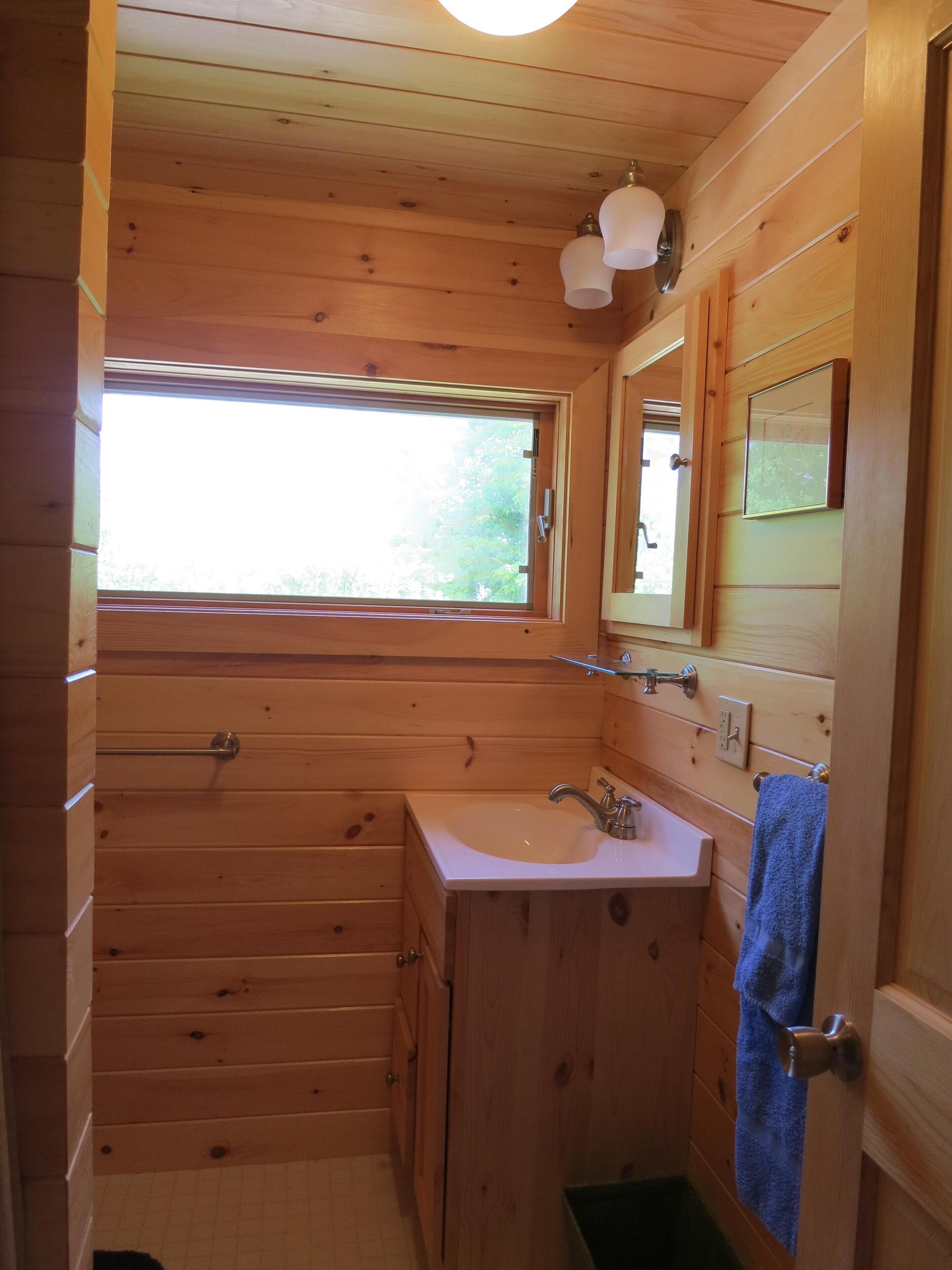 Baño de cabaña de madera con ventana, lavamanos y espejo.