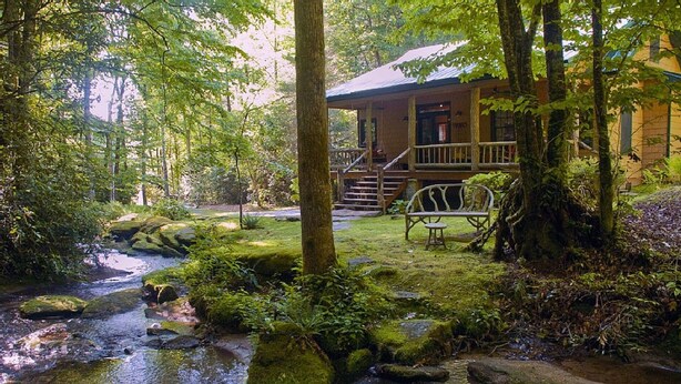 A rustic cabin with a porch and a bench, surrounded by a forest and a small stream.
