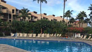 A swimming pool with lounge chairs, palm trees, and a multi-story building in the background. - OCEAN VIEW, LARGE LANAI, STEPS FROM THE BEACH, BLDG 10 (Kihei)