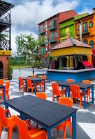 Un espacio al aire libre con mesas y sillas, una parrilla y edificios coloridos al fondo.