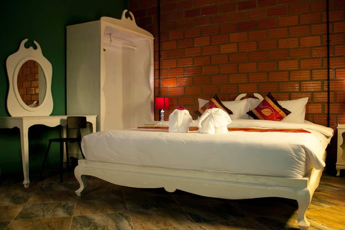 A neatly made bed with white linens and pillows, a white wardrobe, a small table with a lamp, and a brick wall in the background.
