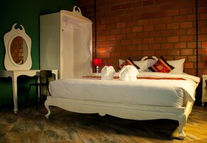 Una cama bien hecha con sábanas y almohadas blancas, un armario blanco, una mesita de noche con una lámpara, y una pared de ladrillo al fondo.