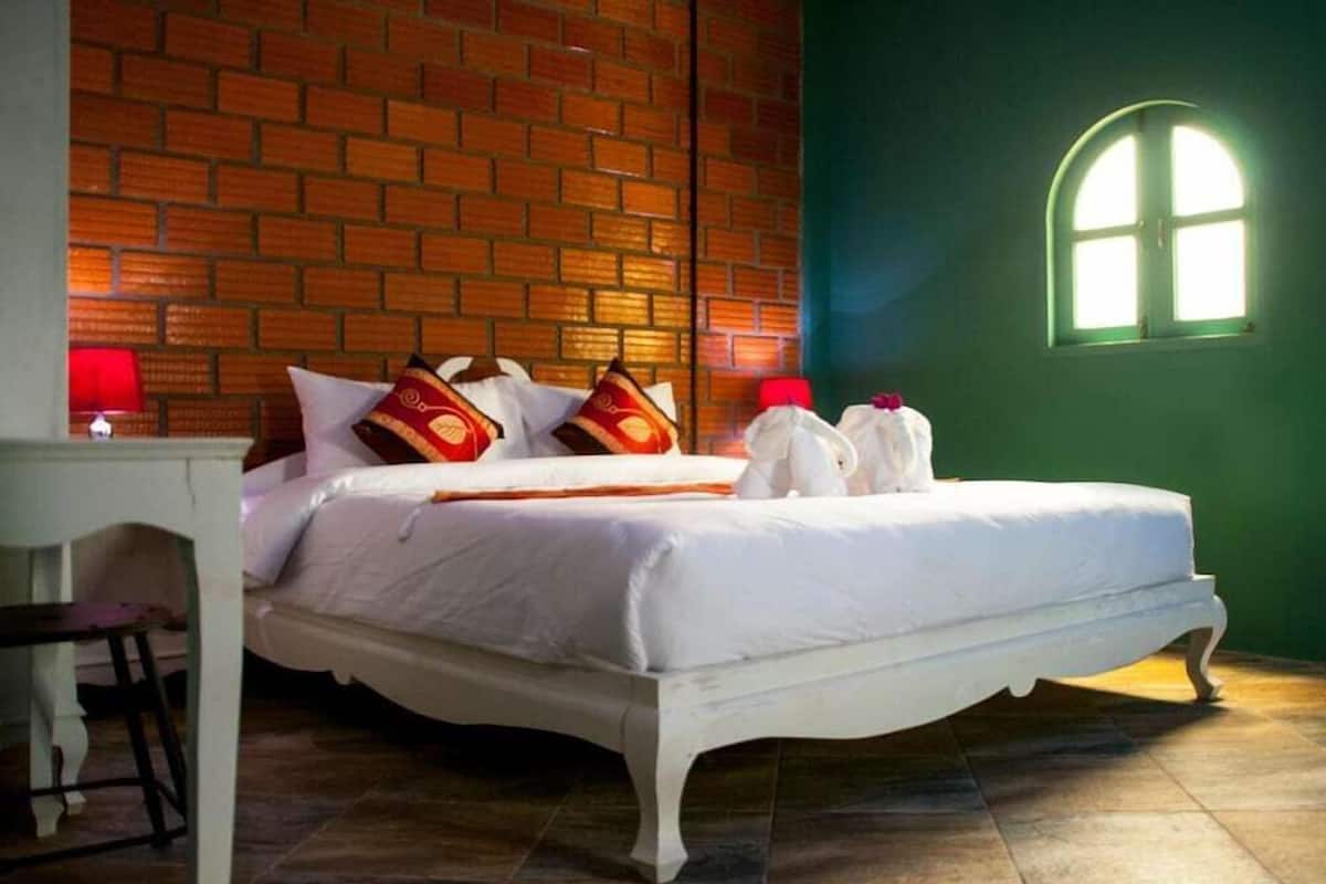 A room with a brick wall, a bed with white bedding, a small table with a red lamp, and a window with a green wall.