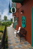 Una terraza con suelo de baldosas, sillas blancas, un banco negro y un edificio rojo con detalles verdes y ventanas circulares.