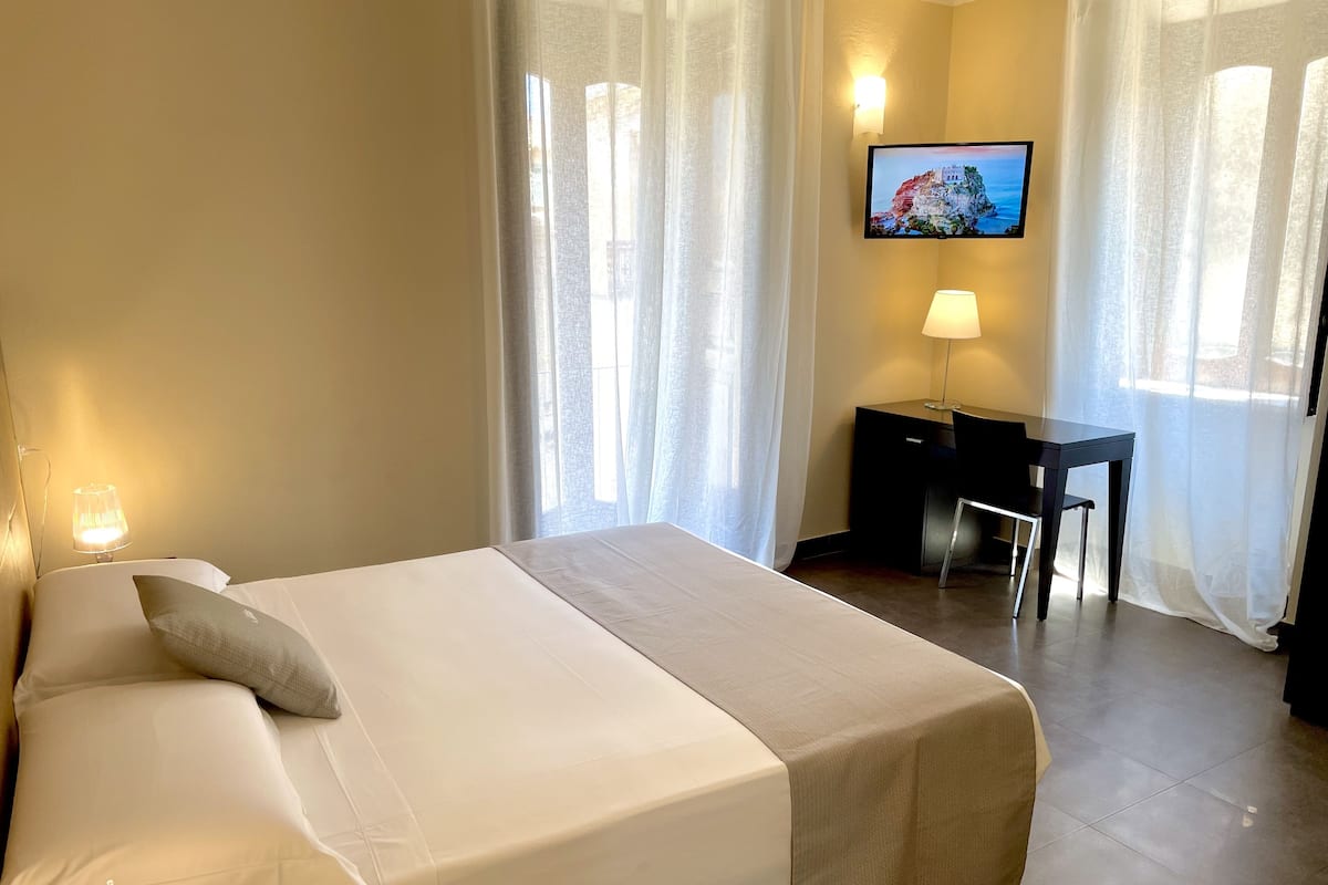 A hotel room with a bed, a desk, a chair, a TV, and a lamp.