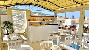 A sunlit outdoor dining area with white chairs and tables, a small kitchen with fruit and beverages, and a view of the cityscape. - Residenza il Barone (Tropea)