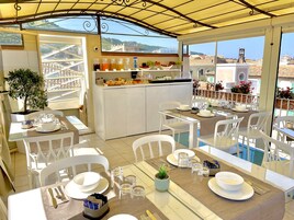 Un área de comedor al aire libre iluminada por el sol, con sillas y mesas blancas, una pequeña cocina con fruta y bebidas, y vistas a la ciudad.