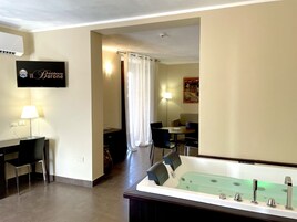 A hotel room with a large bathtub, a dining area with a table and chairs, a TV mounted on the wall, and a painting on the wall.