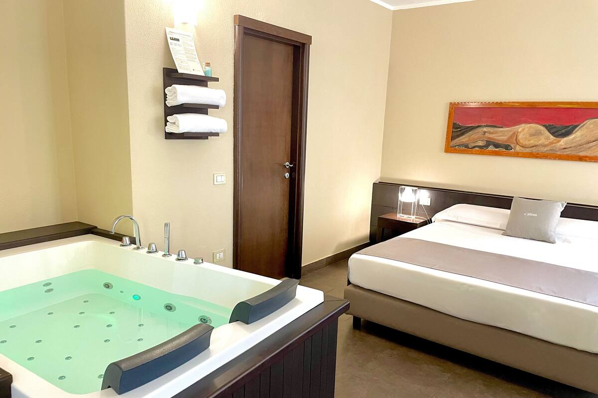 A hotel room with a large bathtub, a bed, a nightstand, and a painting on the wall.