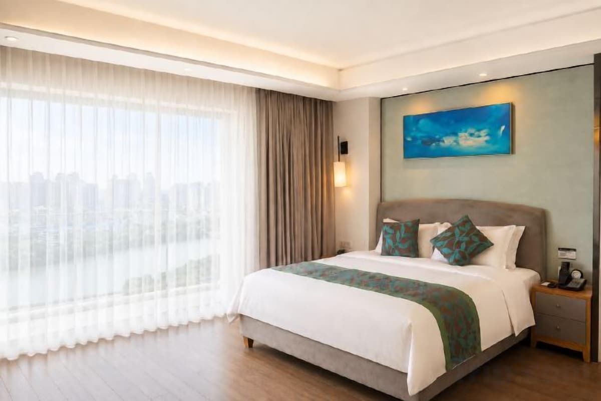 A hotel room with a large bed, a nightstand, a painting on the wall, and a view through the window.