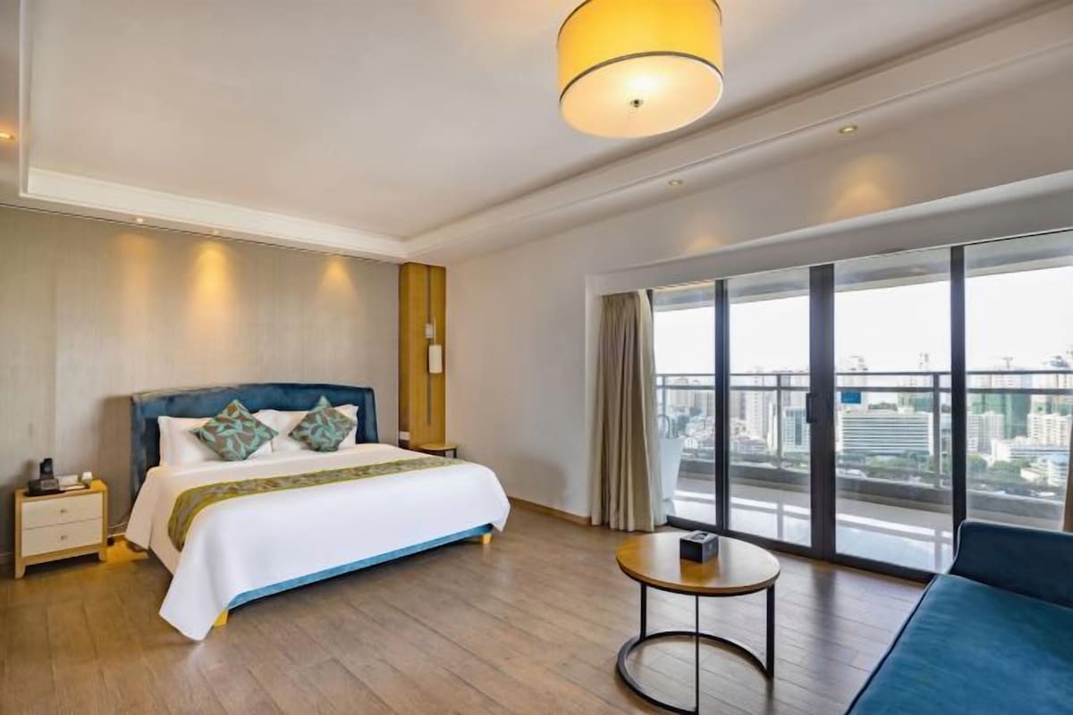A hotel room with a large bed, a round table, and a view of the city through large windows.