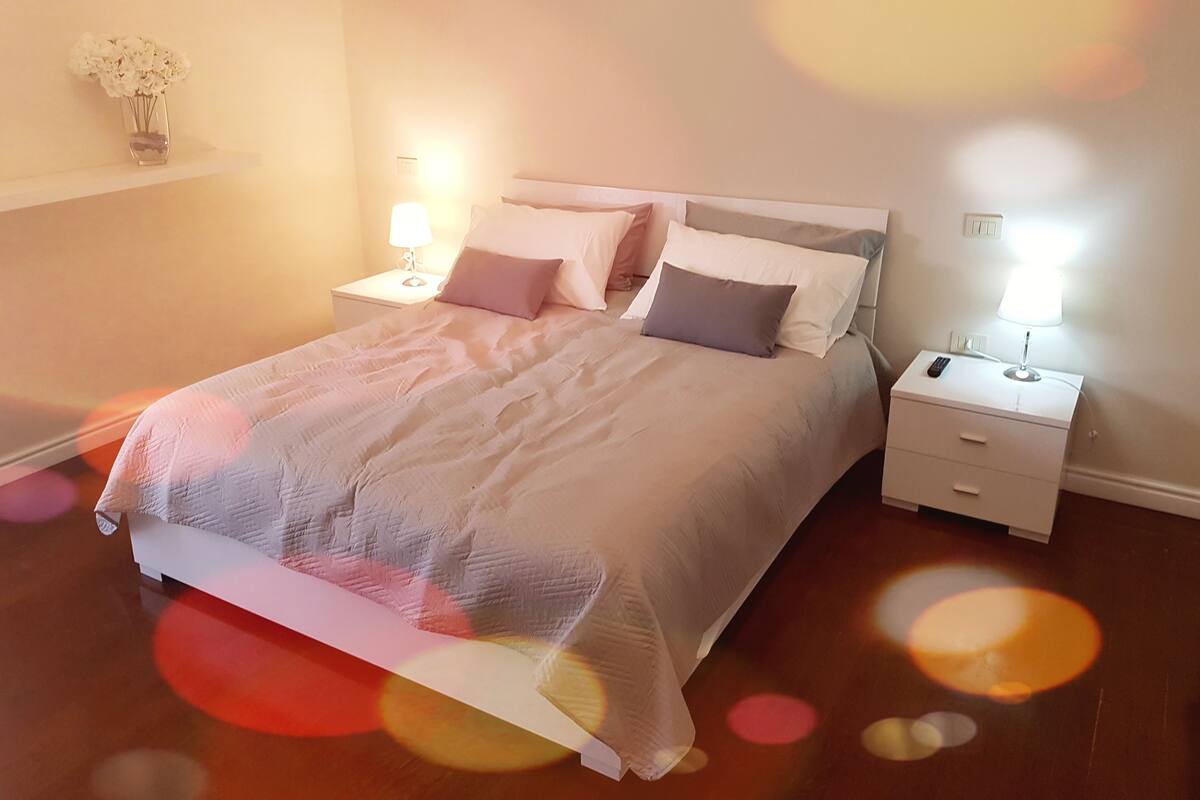 Un lit bien fait, avec une couette blanche et des oreillers, une table de chevet avec une lampe, et une étagère fixée au mur sur laquelle est posé un vase de fleurs.