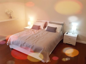 A neatly made bed with a white comforter and pillows, a bedside table with a lamp, and a wall-mounted shelf with a vase of flowers.