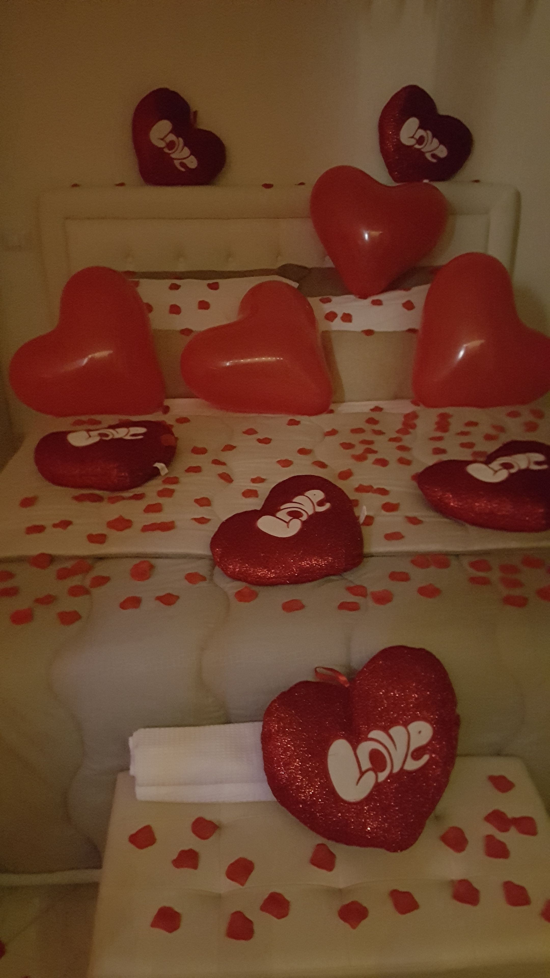 Una cama con múltiples almohadas y una manta, cada una decorada con almohadas con forma de corazón rojo y pétalos de rosa rojos.