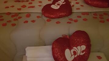 Una cama con múltiples almohadas y una manta, cada una decorada con almohadas con forma de corazón rojo y pétalos de rosa rojos.