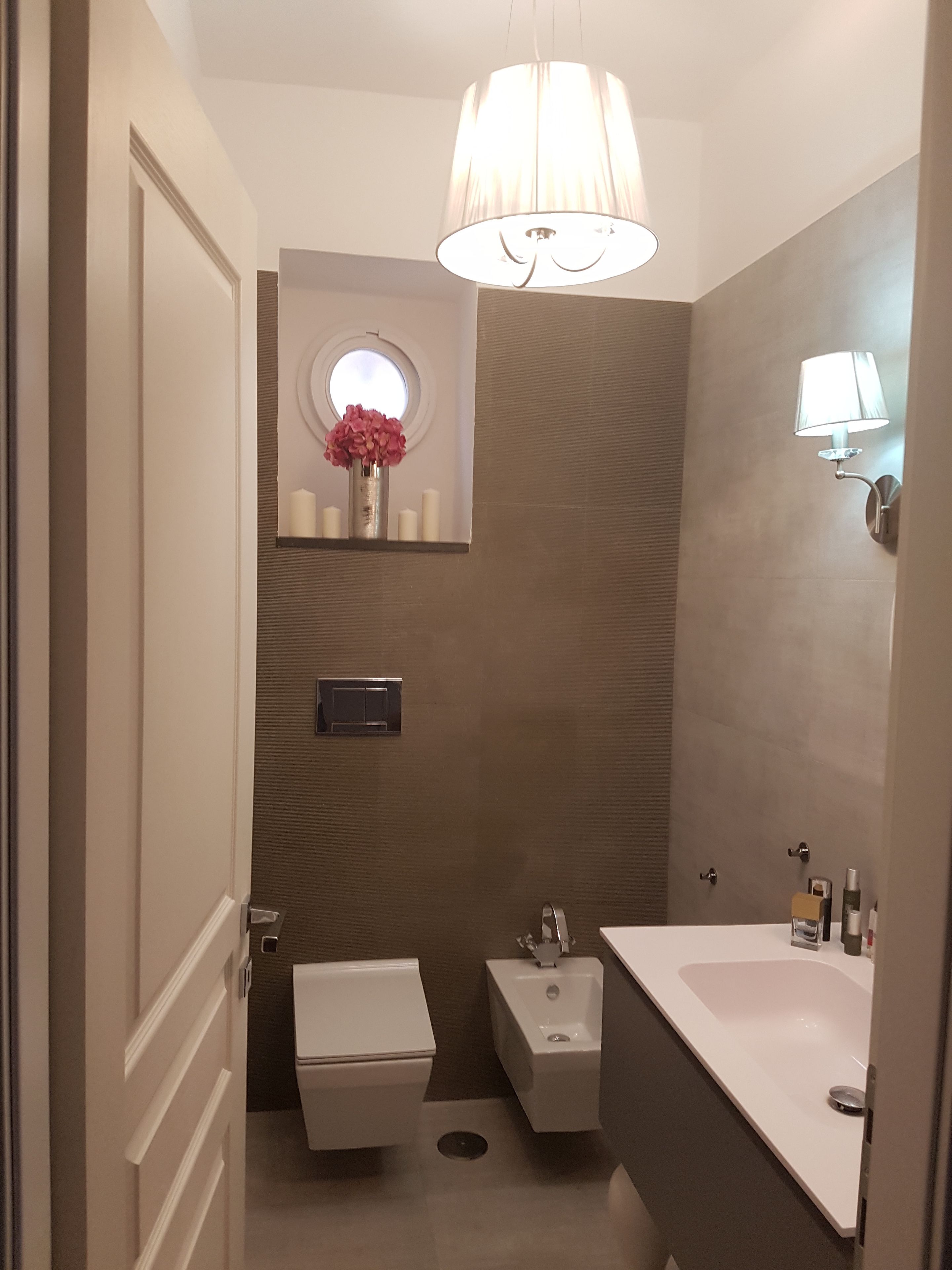 A modern bathroom with a toilet, bidet, and sink. There is a mirror with a round window above the toilet, and a wall-mounted light fixture.