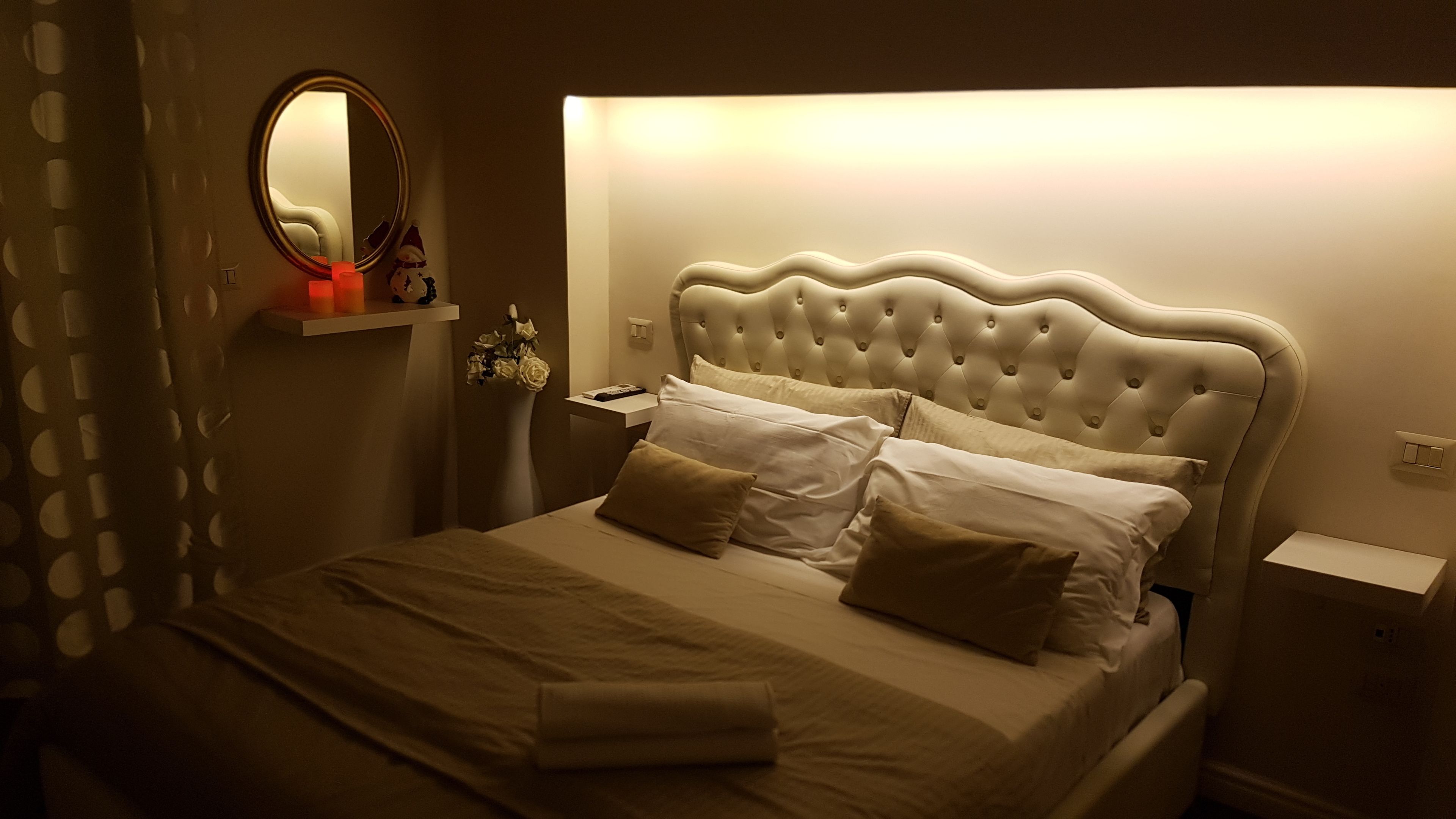 A neatly made bed with a tufted headboard, a bedside table with a lamp, and a wall-mounted mirror.