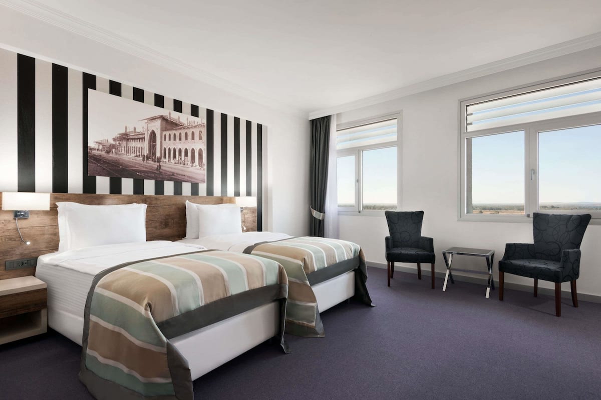 A hotel room with two beds, a desk, two chairs, a large window, and a wall-mounted picture.