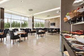 Un comedor amplio con varias mesas y sillas, un bufé con diferentes opciones de comida y ventanas grandes con vistas a la vegetación exterior.