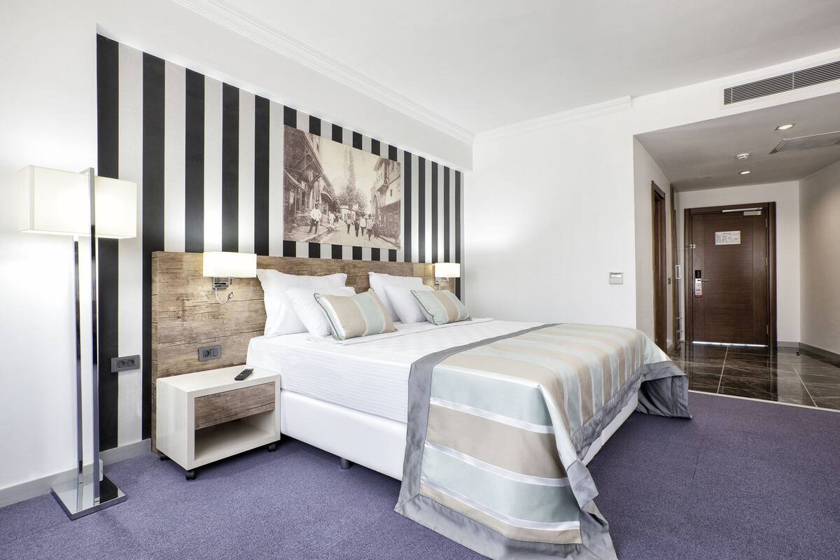 A modern hotel room with a large bed, a nightstand, a wall-mounted lamp, and a classic painting.