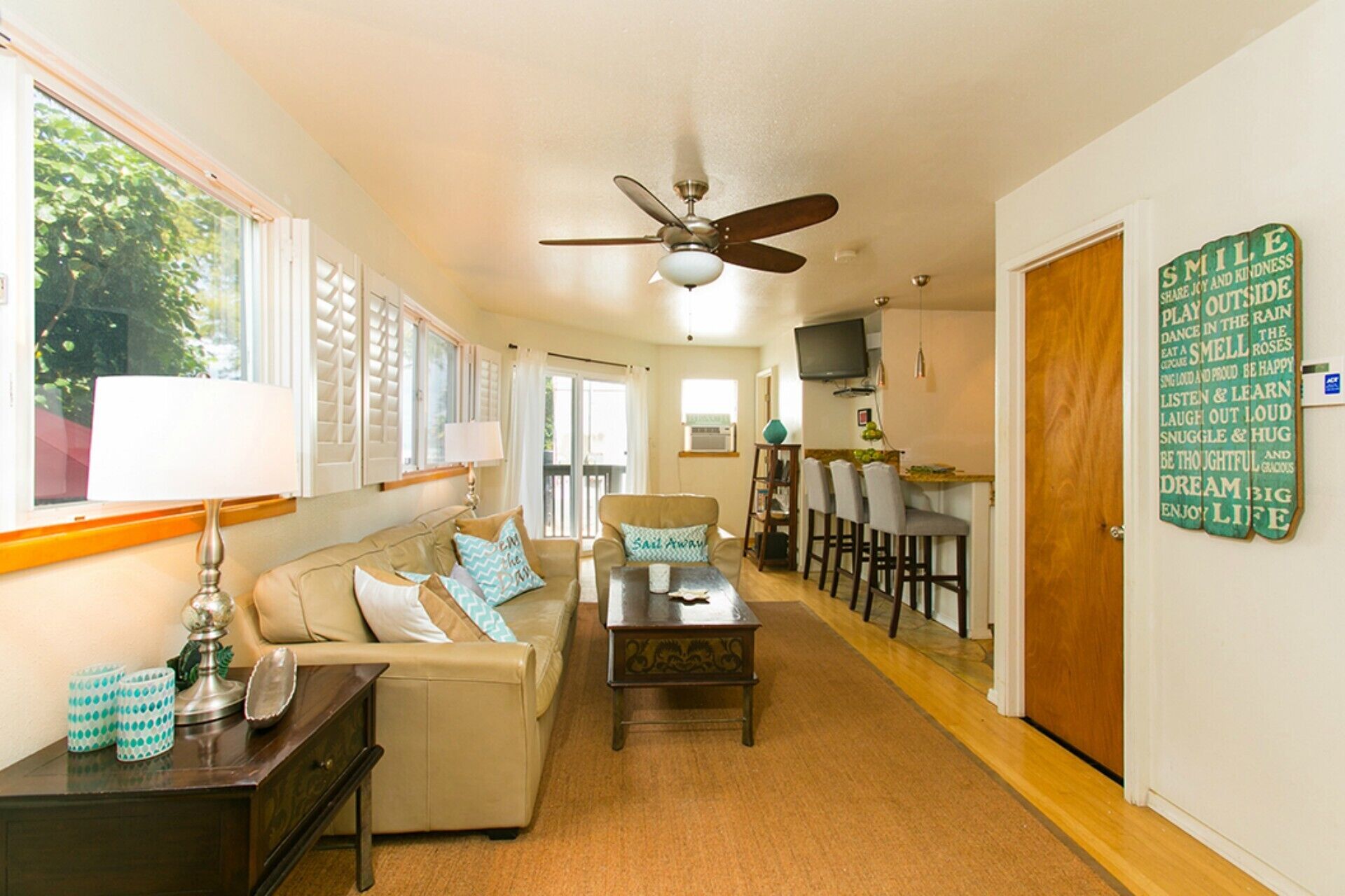 A spacious living room with a sofa, coffee table, and a ceiling fan. There is a dining area with chairs and a table, and a kitchen area with a TV.