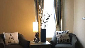 Ein Zimmer mit zwei Sesseln, einem kleinen Tisch, einer Lampe und einem fenster mit Vorhängen.