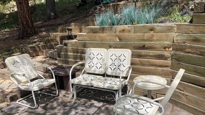 A patio area with a wooden bench, two white chairs with intricate back designs, and a small round table.