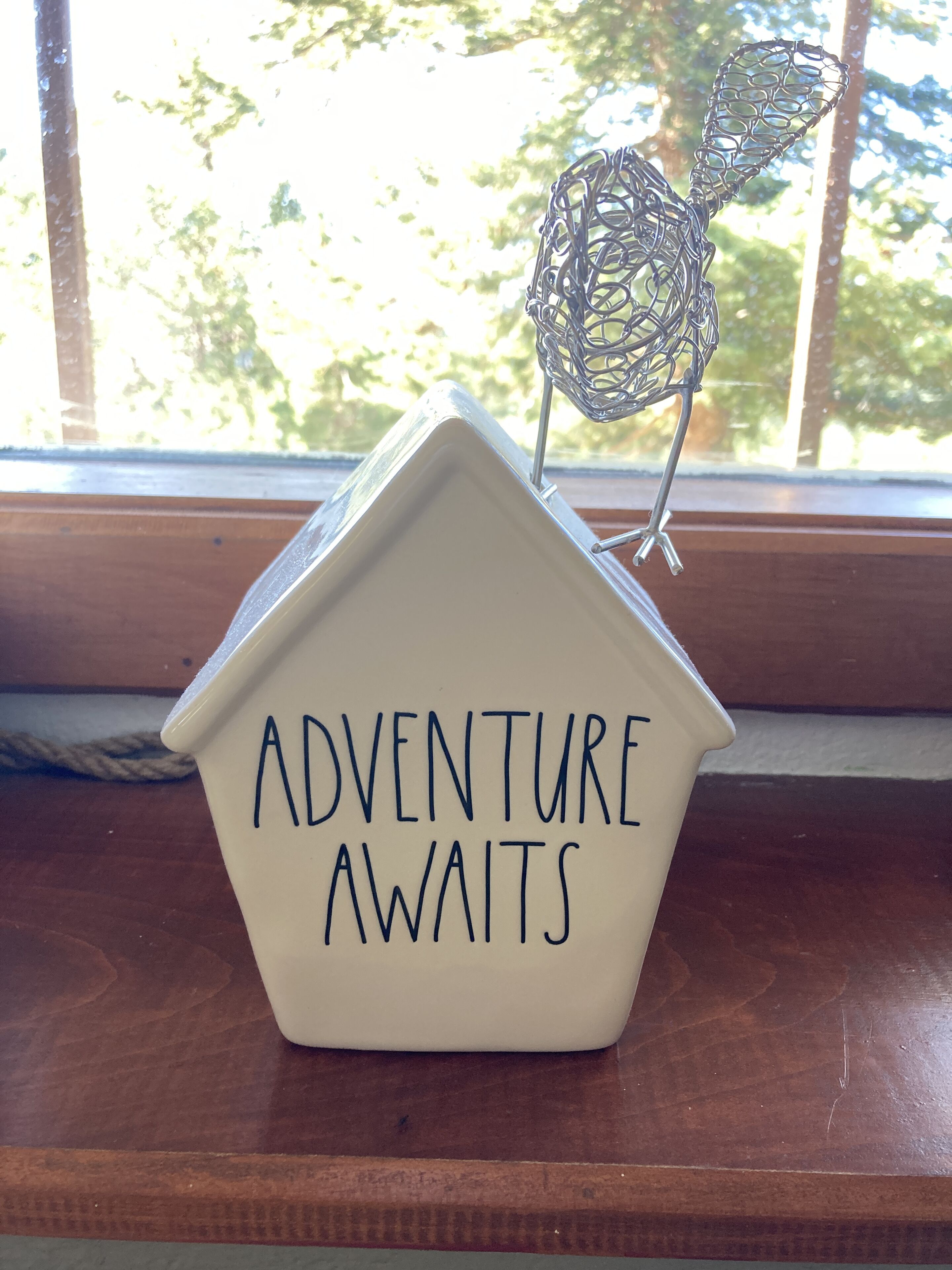 A ceramic house-shaped container with the inscription "ADVENTURE AWAITS" and a metal wire sculpture of a bird.