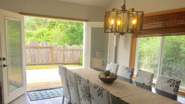 A dining area with a table set for six, chairs with floral upholstery, a chandelier, and a view of a wooden deck and greenery outside. - No Host Fees! Lovely, well equipped, dog friendly home, 3 blocks from the beach! (Rockaway Beach)