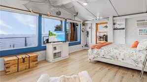 A room with a large window overlooking the sea, a bed with floral bedding, a wooden chest, and a small desk with a lamp. - The Boat House · Riverfront · Heated Pool & Spa · Tiki Bar · Sleeps 10 (Cape Coral)